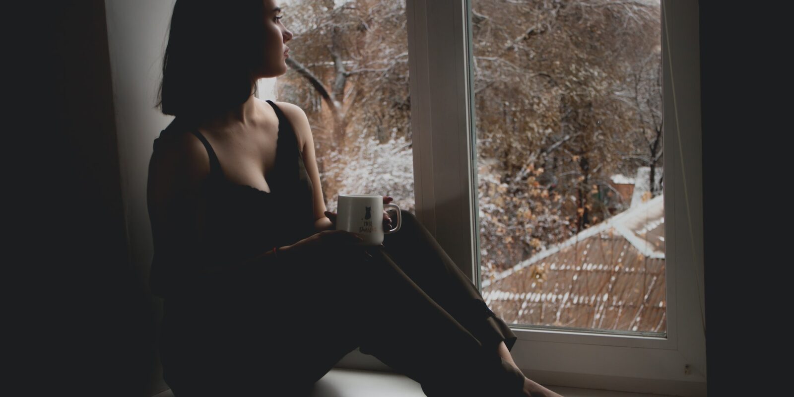 A sad woman sitting near a window with a cup of warm tea that's useful in heart breaks